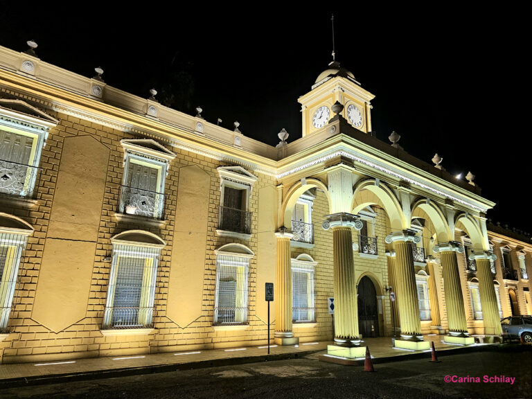 Das Rathaus von Santa Ana in El Salvador bei Nacht, ein gelbes Gebäude mit weißen Säulen, Bögen und einem Uhrturm.