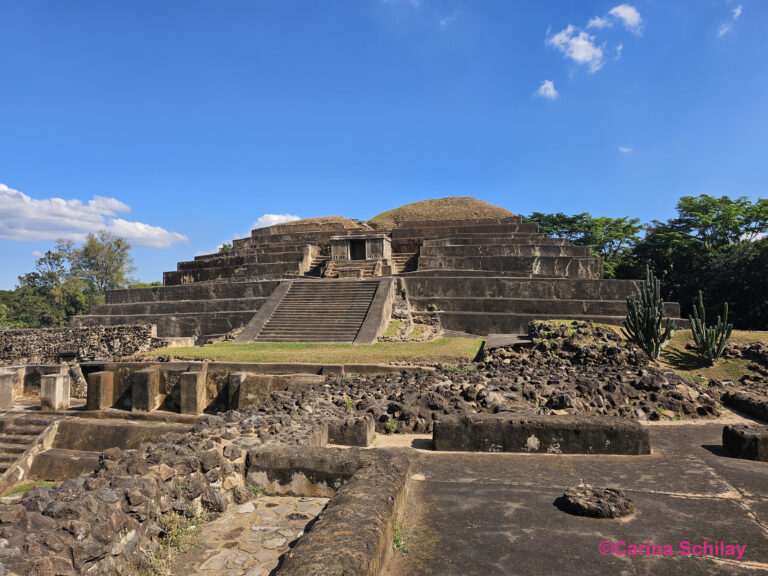 Eine Ansicht der beeindruckenden Ruinen von Tazumal mit einer majestätischen Pyramide, umgeben von grüner Flora unter einem klaren blauen Himmel.