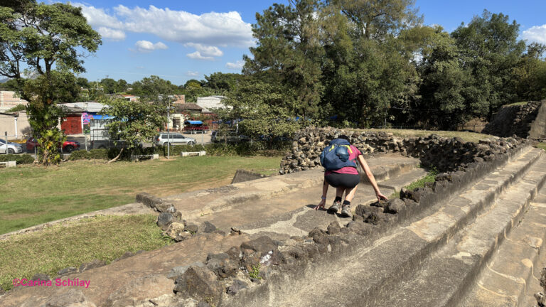Eine Reisende erkundet die Steinstrukturen der Tazumal-Ruinen inmitten einer üppigen grünen Landschaft.