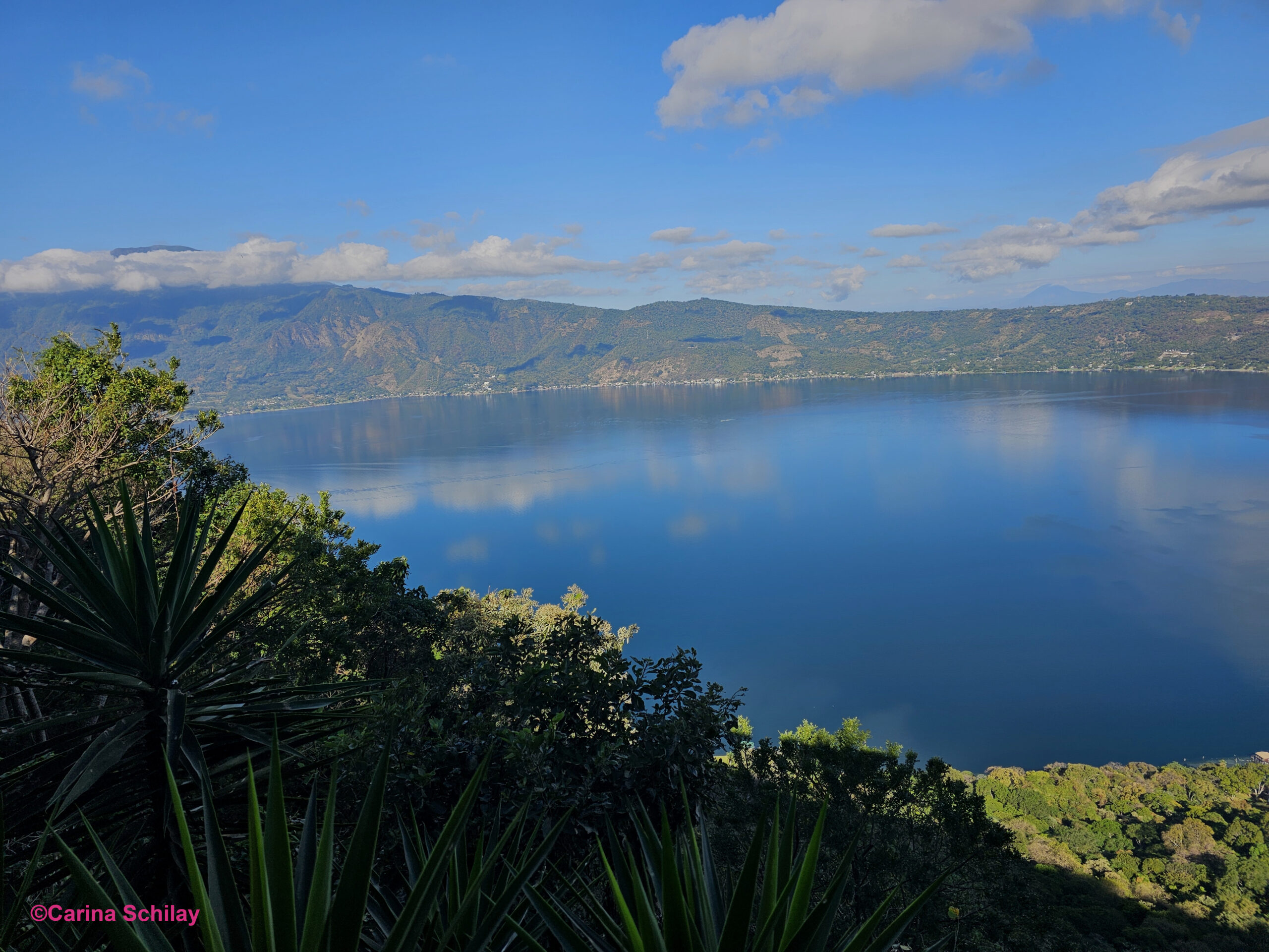 Blick auf die idyllische linke Seite des Lago de Coatepeque in El Salvador, umgeben von üppigem Grün und einem klaren blauen Himmel.