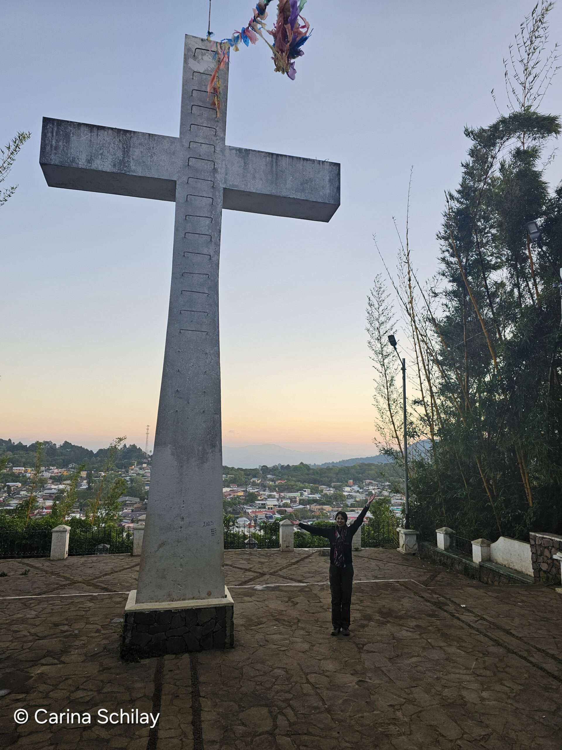 Person steht mit ausgebreiteten Armen vor dem beeindruckenden Gipfelkreuz ‘El Mirador’ in Apaneca, El Salvador, während bunte Bänder im Wind flattern und im Hintergrund die malerische Stadtansicht zu sehen ist.