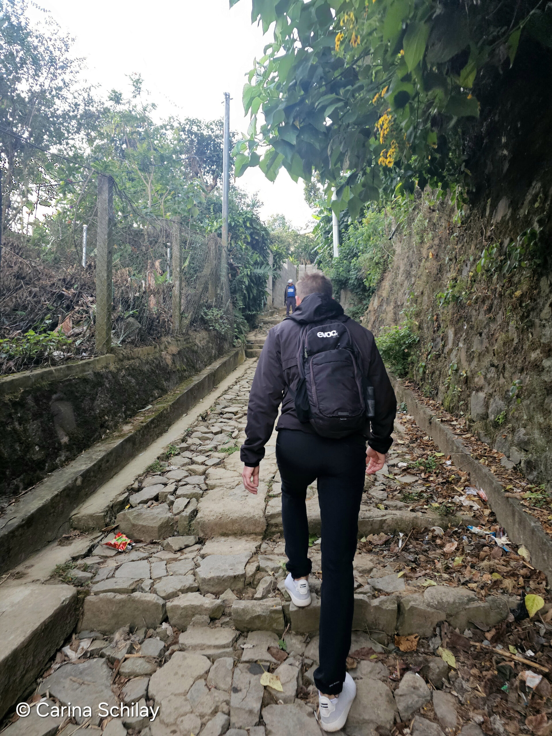 Ein Wanderer, erkennbar von hinten, geht einen steinigen Weg entlang, umgeben von üppigem Grün und blühenden Pflanzen an der Ruta de Flores in El Salvador.