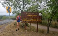 Person steht vor einem hölzernen Schild des Nationalmonuments Somoto-Schlucht, umgeben von einer grünen Landschaft mit Straße und Bäumen im Hintergrund.